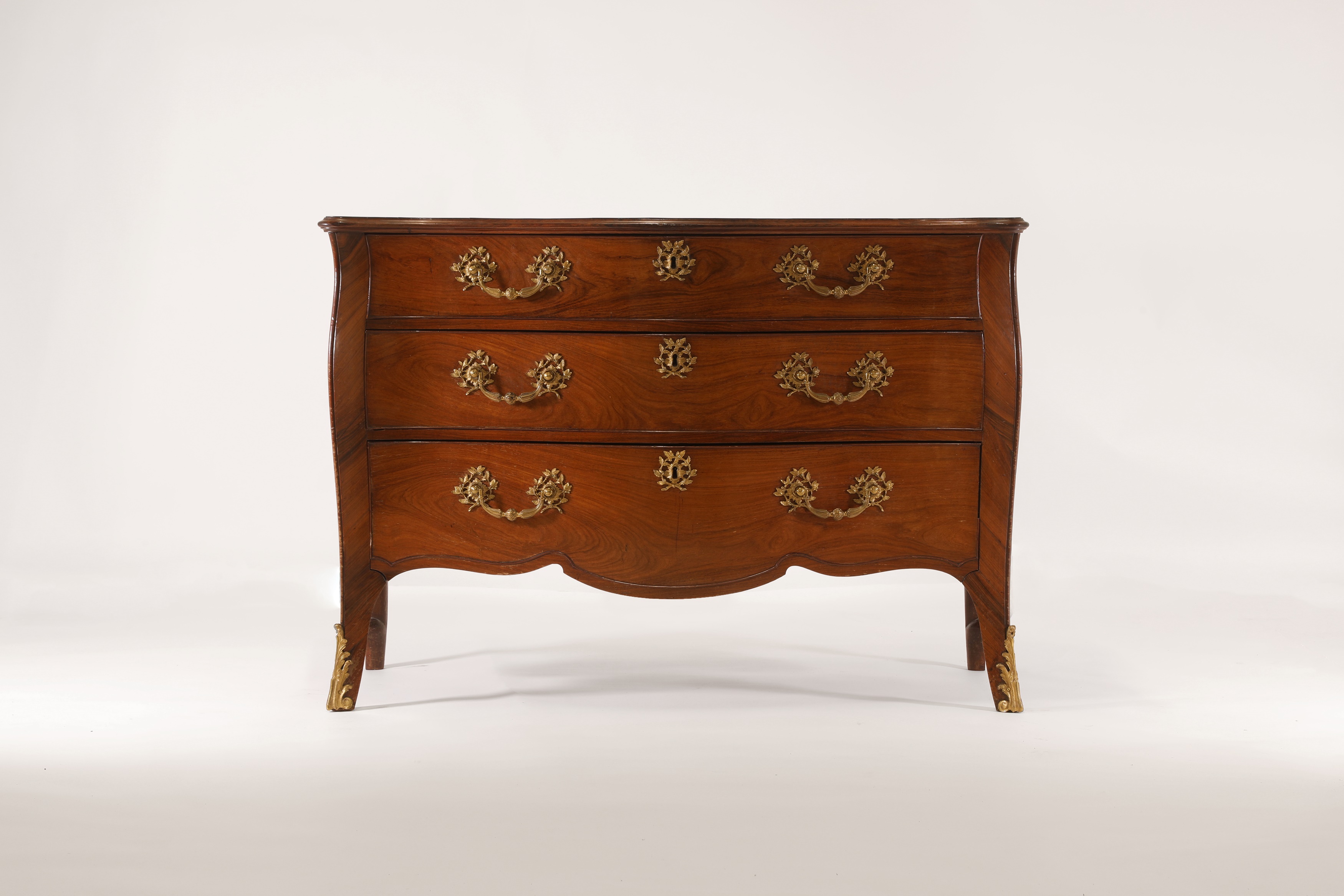 A George III padouk and kingwood commode attributed to John Cobb, c.1765, of serpentine outline with ormolu mounts, the matched-veneered top with a crossbanded moulded edge above three long cockbeaded drawers mounted with laurel wreath and reeded bail handles, with conforming escutcheons and carry handles to the sides above a wavy edge apron, on splayed forelegs terminating in foliate-cast sabots, 125cm wide 65cm deep 88cm high (£30,000-50,000)